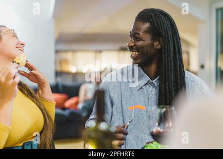 L'uomo africano di mezza età con greggi in camicia di denim sorride mentre cena con gli amici - una donna caucasica curvy ride e mangia nachos al bordo o Foto Stock