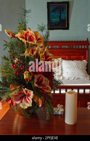 Un coloratissimo allestimento di fiori di Natale e un piccolo omaggio sono esposti su un tavolo da letto con un letto coperto di biancheria bianca sullo sfondo del 187 Foto Stock