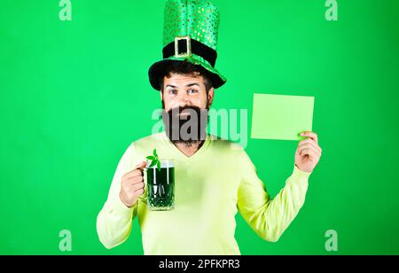 Giornata dei Patricks. Uomo barbuto in leprechaun cappello con birra verde tiene la scheda pubblicitaria. Simboli del giorno di San Patrizio. Tradizione irlandese. Patricks Day uomo Foto Stock