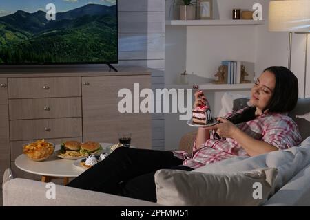 Donna felice in sovrappeso che mangia la torta mentre guarda la TV sul divano a casa Foto Stock