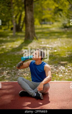Un bell'uomo è seduto su una pista sportiva nel parco, stanco, dopo una giornata di sole. È acqua potabile Foto Stock