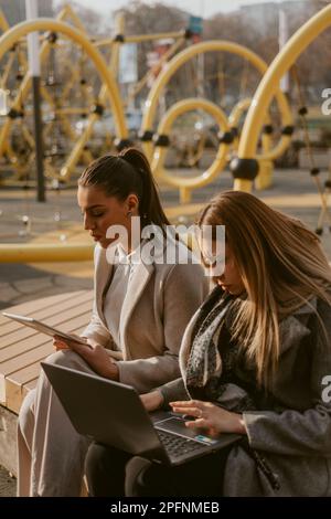 Due donne in viaggio d'affari che lavorano sul loro notebook e tablet mentre si siedono sulla panca di legno del parco Foto Stock