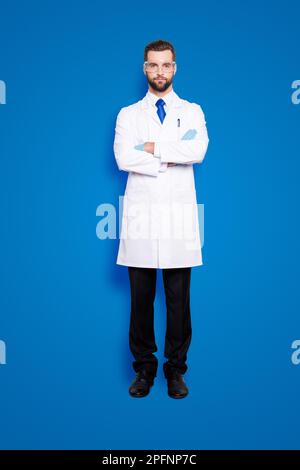 Ritratto integrale di elegante e bello scienziato con stoppia in abito bianco con cravatta blu con le braccia incrociate guardando la fotocamera Foto Stock