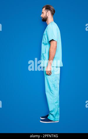 Bottoncino a figura intera, mezza faccia vista laterale ritratto di bell'uomo attraente con setole, acconciatura in tuta da laboratorio blu, isolato su sfondo grigio Foto Stock