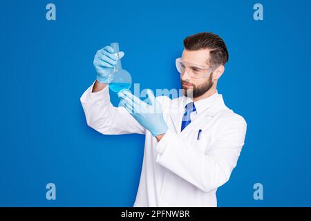 Ritratto di scienziato occupato concentrato con stoppia in bianco camice da laboratorio, guanti esaminare, guardando il pallone con liquido multicolore nel braccio Foto Stock