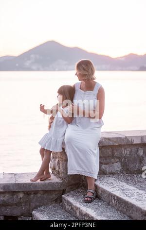 Madre millenaria incinta siede con figlia al tramonto sul mare. La donna di mezza età abbraccia una bambina. Il viaggio del genitore singolo. Gravidanza sana Foto Stock