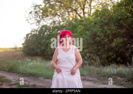 Donna diversità luminosa con ritratto dei capelli rosa. La ragazza si è schiacciata su una strada forestale verso il tramonto. Ragazza Childfree, viaggio nel fine settimana. Individuale, bol Foto Stock