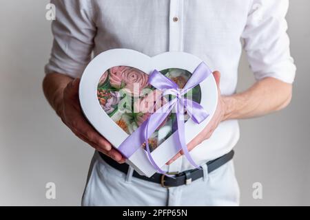 L'uomo senza volto tiene in mano la forma scatolata del cuore di fiori dolci marshmallow. Regalo con nastro viola. Delicate rose senza zucchero su sfondo chiaro. SM Foto Stock