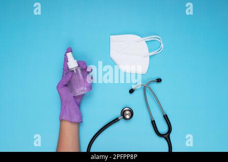 Lilla medico guanto mano che tiene spray disinfettante, maschera chirurgica bianca e stetoscopio su sfondo azzurro Foto Stock