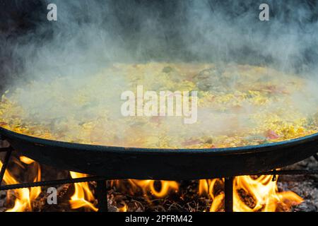 Una grande paella che cucina sopra il fuoco con un fondo di fumé Foto Stock