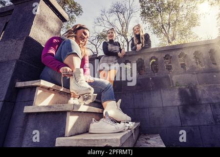 Amici diversi che chiacchierano in un parco, uno sulle scale con pattini a rotelle, due su un balcone, in un tiro basso-angolo con sole flare - giovani lifestyle co Foto Stock