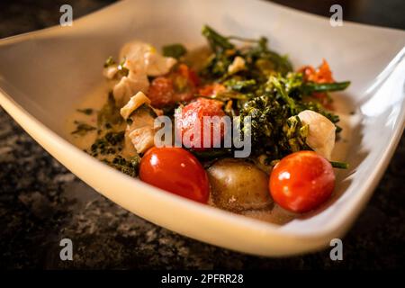 Una deliziosa zuppa tailandese con latte di cocco, pollo, pomodori ciliegini, broccoli e patate, servito in una ciotola bianca su uno sfondo grigio a motivi geometrici. Foto Stock