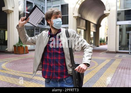 Un lavoratore di ufficio che indossa una maschera medica che è stato licenziato dal suo lavoro colpisce al di fuori di un edificio di ufficio. Concetto di disoccupazione e licenziamenti Foto Stock