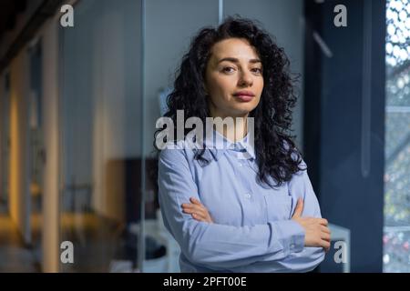 Ritratto di boss donna serio all'interno dell'ufficio aziendale, donna d'affari ha attraversato le braccia guardando concentrato alla macchina fotografica, indossando una camicia, donna ispanica soddisfatta e di successo con capelli ricci. Foto Stock
