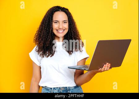 Foto di una donna brasiliana o ispanica con capelli ricci, che indossa una t-shirt bianca di base, che tiene in mano un computer portatile aperto, guarda la fotocamera con sorriso, in piedi su uno sfondo giallo isolato Foto Stock