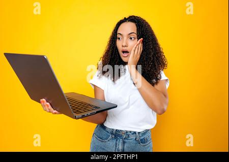 Scioccato emotivo brasiliano o donna ispanica con capelli ricci, vestito in abbigliamento casual, tenendo un notebook aperto in mano, guardando lo schermo del notebook in sorpresa, in piedi su sfondo giallo isolato Foto Stock
