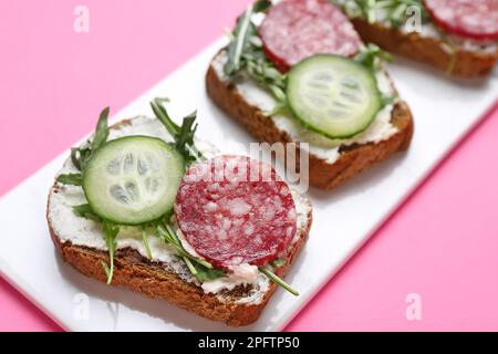 Piatto di gustosi sandwich con formaggio cremoso, salame, cetriolo e rucola su sfondo colorato, primo piano Foto Stock