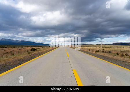 Una lunga strada diritta con linee gialle che conducono verso le montagne che attraversano il deserto di Atacama in Cile. Sole con nuvole nel cielo blu. Foto Stock