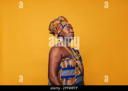 Donna matura che indossa un abbigliamento tradizionale africano su uno sfondo giallo. Donna nera sicura vestita in tessuto colorato Kente e jewe dorato Foto Stock