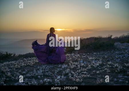 Tramonto viola abito donna montagne. Ascesa del mistico. tramonto sulle nuvole con una ragazza in un lungo vestito viola. Nel prato c'è un prato Foto Stock