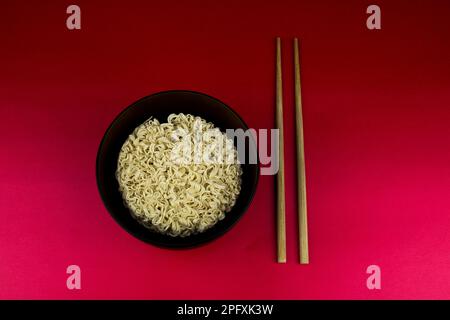 Blocco di spaghetti istantanei in una ciotola di legno su un tavolo rosso con bastoncini Foto Stock