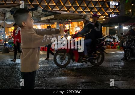 Un ragazzo si alza con le braccia tese sulla strada al mercato notturno di Dalat, in Vietnam. Foto Stock