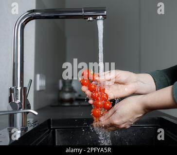 Pomodori ciliegini freschi rossi sul ramo sotto il flusso d'acqua nel lavandino, donna in cucina lavando le verdure prima di cucinare, senza volto Foto Stock