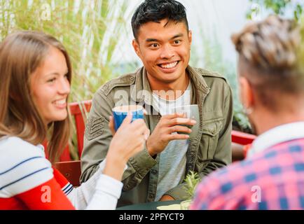 Allegro uomo e donna multirazziale con tazze di caffè sorridenti e ascoltando gli amici mentre si siede al tavolo nel caffè all'aperto Foto Stock