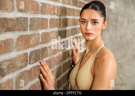 Ballerina giovane e attraente con un'acconciatura da bun che guarda la macchina fotografica mentre si trova vicino a una parete in mattoni in un moderno studio luminoso Foto Stock