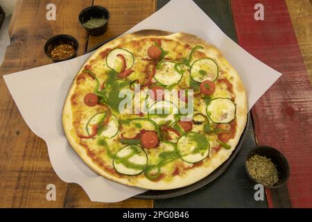 Una meravigliosa pizza a base di crosta media e sottile con vari tipi di verdure fresche, pomodori ciliegini, pesto e pomodoro su un tavolo di legno colorato Foto Stock