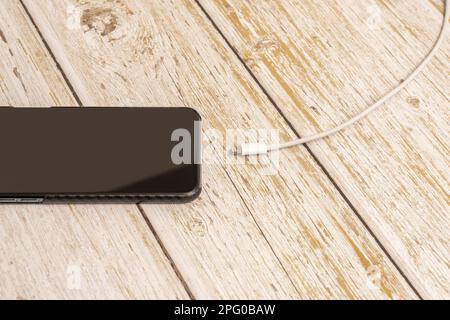 Telefono con custodia in fibra di carbonio e cavo di ricarica bianco scollegato su un tavolo di legno Foto Stock