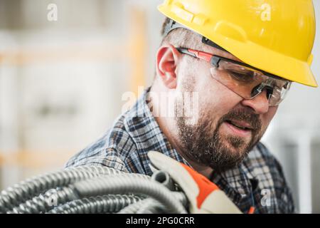 Caucasico professionista lavoratore di elettrico Contraente nel suo 30s con condotto elettrico. Indossare il cappello giallo duro e gli occhiali di protezione. Foto Stock