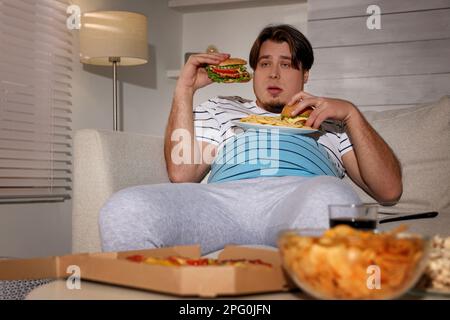 Uomo sovrappeso con piatto di hamburger e patatine fritte sul divano a casa Foto Stock