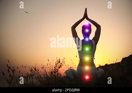 Donna meditando all'aperto al tramonto, vista posteriore. Schema di sette chakra, illustrazione Foto Stock