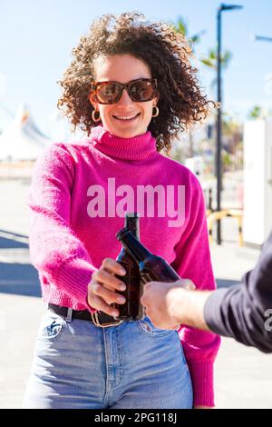 Capelli ricci giovane donna caucasica fare un brindisi con un coltivatore in strada tenendo due bottiglie di birra in una giornata di sole. Foto Stock