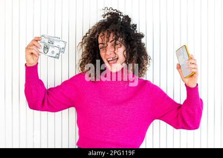 Giovane donna caucasica con capelli ricci è felice, mentre tiene con una mano circa 50 dollari bollette e uno smartphone in altra mano di fronte a un muro. Foto Stock