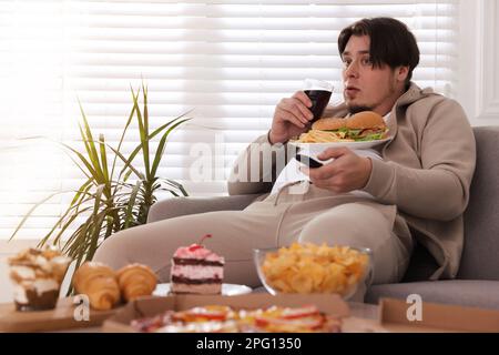 Uomo in sovrappeso con piatto di hamburger e patatine fritte guardando la TV sul divano a casa Foto Stock