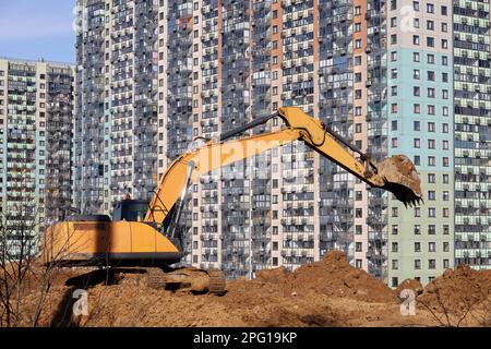 L'escavatore cingolato scarta la terra con una benna sullo sfondo di nuovi edifici residenziali. Lavori di movimento terra, industria di scavo e di costruzione Foto Stock