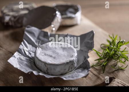 Confezioni di camembert con uno di essi aperto accanto al rosmarino su un asse di legno. Foto Stock