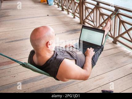 Lavorando a distanza sul suo computer in si Phan Don, Laos meridionale, mentre si viaggia per il mondo, rilassarsi su un balcone pensione in legno che si affaccia sul Mekon Foto Stock