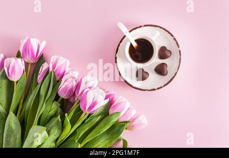 una tazza di caffè espresso si trova su un piattino con cioccolatini a forma di cuore e un bouquet di tulipani lilla. sfondo festivo. vista dall'alto Foto Stock