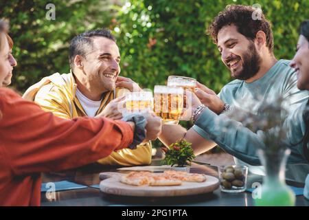 Un gruppo di cinque amici seduti a un tavolo in giardino, alzando tazze di birra per brindare. Una deliziosa pizza è su un tagliere di fronte a loro - focus o Foto Stock