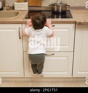 Il bambino salite su una stufa elettrica calda nella cucina domestica. Un bambino piccolo tocca la superficie della stufa con la mano al rischio di ottenere b Foto Stock