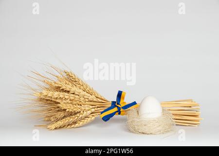 Spikelets di grano legato con un nastro della bandiera dell'Ucraina e un uovo bianco di pollo in un nido su uno sfondo bianco, vacanza Foto Stock