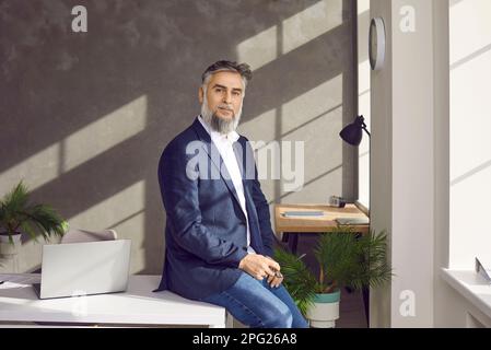 Uomo d'affari maturo seduto sul tavolo del suo posto di lavoro in ufficio, guardando sicuro di fotocamera. Foto Stock