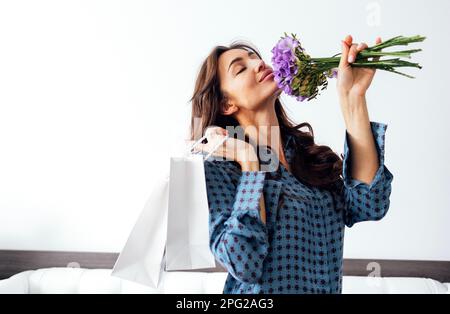 Giovane bella donna chiuse gli occhi e odora bouquet di fiori con piacere. Donna acquirente in abbigliamento casual che tiene due shopping di carta bianca Foto Stock