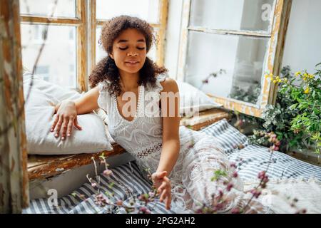 Bella donna afroamericana sorridente in pizzo bianco vestito toccando ramo fiorito. Bella giovane ragazza pelle scura si trova sul materasso e cuscini vicino ope Foto Stock