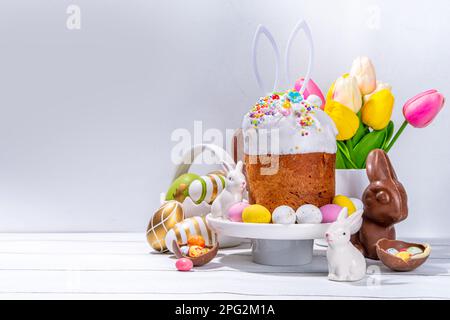 Gustoso torta di Pasqua glassato con decorazione di zucchero, cestino di uova colorate, conigli in ceramica e fiore di primavera sul tavolo da cucina bianco, spazio copia. Buona Pasqua ho Foto Stock