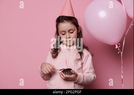 Ragazza piccola felice in cappello rosa festa, celebrando il suo compleanno, mangiando delizioso cupcake al cioccolato. Bambini. Eventi festivi Foto Stock