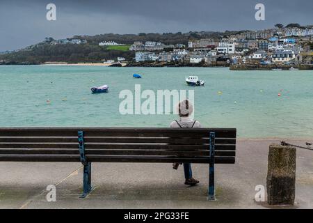 Una donna seduta da sola su una panchina che si affaccia sul porto in una fredda giornata umida a St Ives in Cornovaglia in Inghilterra nel Regno Unito. Foto Stock